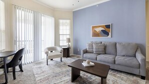 Una sala de estar con un sofá gris, una mesita, una silla y un centro de café. Hay una ventana con persianas y un cuadro en la pared.