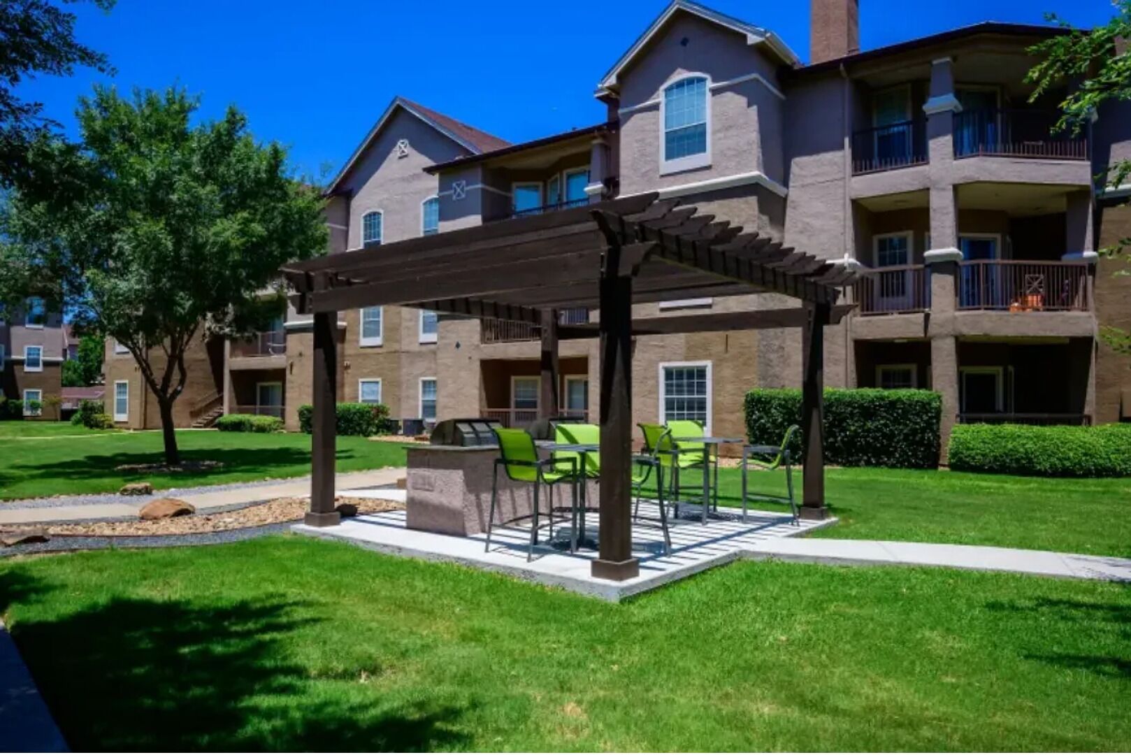 A covered outdoor seating area with tables and chairs, surrounded by a well-maintained lawn and a multi-story building in the background.