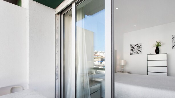 A modern hotel room with a sliding glass door, a white bed, a bedside table, a lamp, and a view of the cityscape. - Natalina T1 Apartment with Balcony and Wi-Fi (Nazaré)
