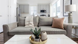 A modern living room with a grey sofa, a coffee table with decorative items, and a kitchen in the background. - Landing | Amazing 2BD, Gym (Phoenix)