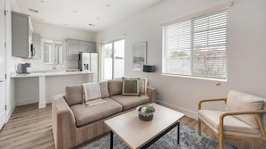 Ein modernes Wohnzimmer mit einer Couch, einem Couchtisch und einem Sessel. Es gibt eine Küchenzeile mit Schränken und einem Kühlschrank.