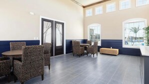A spacious indoor area with a high ceiling, a seating arrangement with wicker chairs and a round table, and large windows providing natural light. - Landing | Stylish 3BD, Gym, Pool (Cocoa)