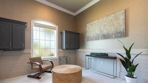 A spacious room with a large window, dark cabinets, a woven ottoman, a console table, and a potted plant. - Landing | Stylish 3BD, Gym, Pool (Cocoa)