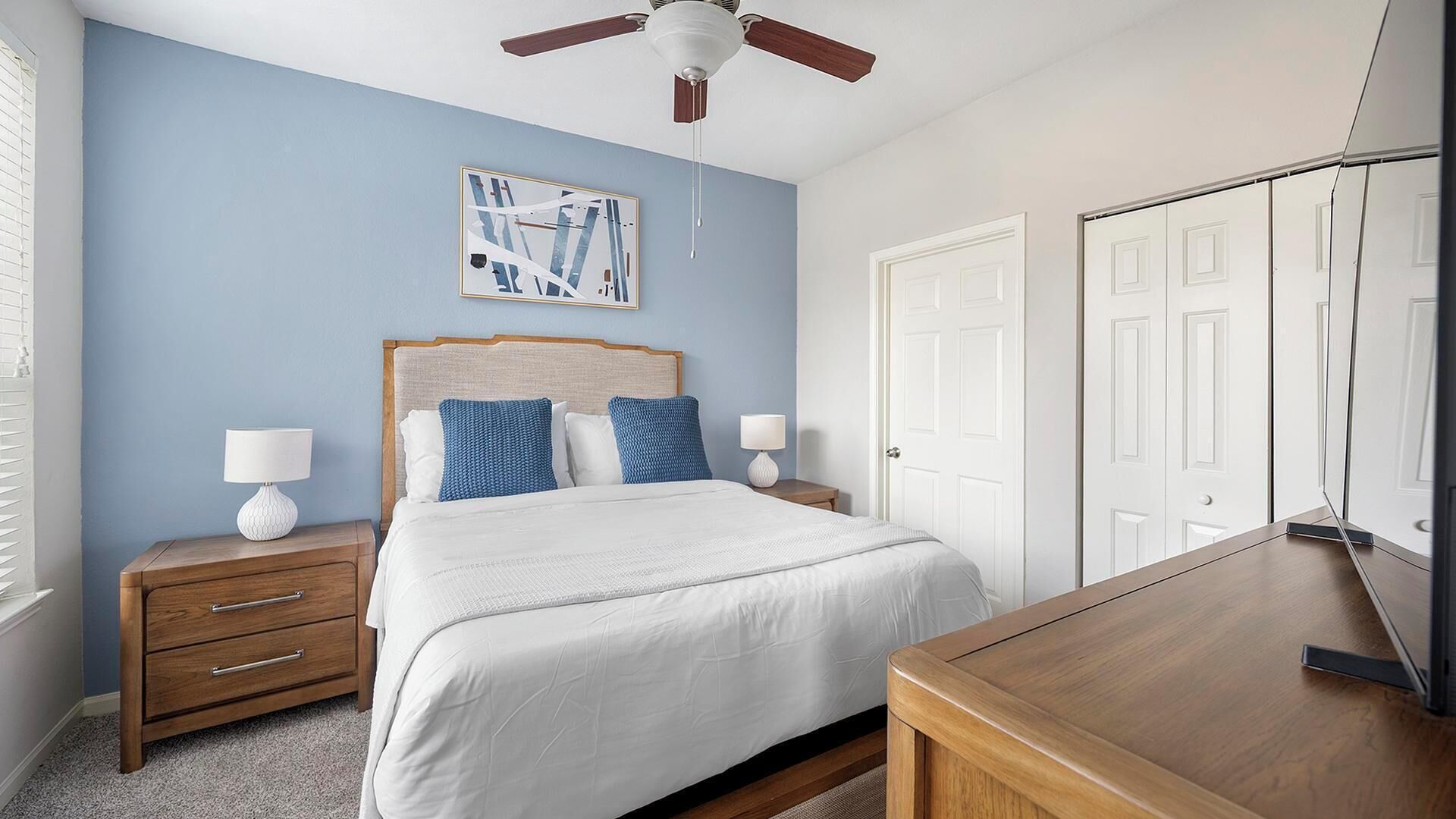 Une chambre à coucher comprenant un lit, des tables de chevet, un ventilateur de plafond, un bureau et une porte.