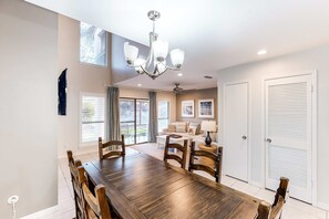 A dining area with a wooden table and chairs, a chandelier, and a view of a living room with a sofa and a ceiling fan. - Exclusive 2BR condo w/ pools & hot tub (Miramar Beach)