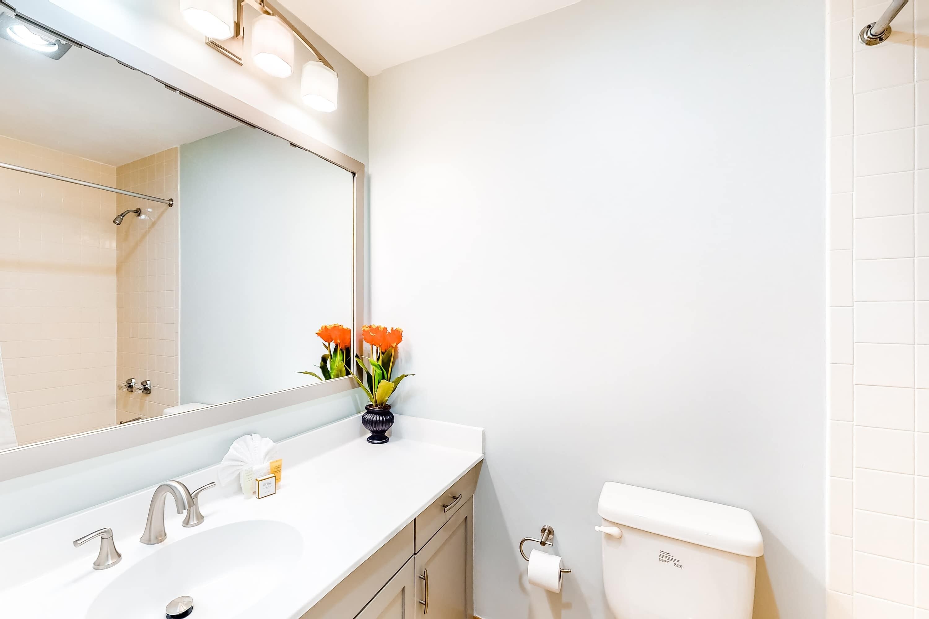 A bathroom with a white sink, two faucets, a mirror, a toilet, and a vase with flowers.