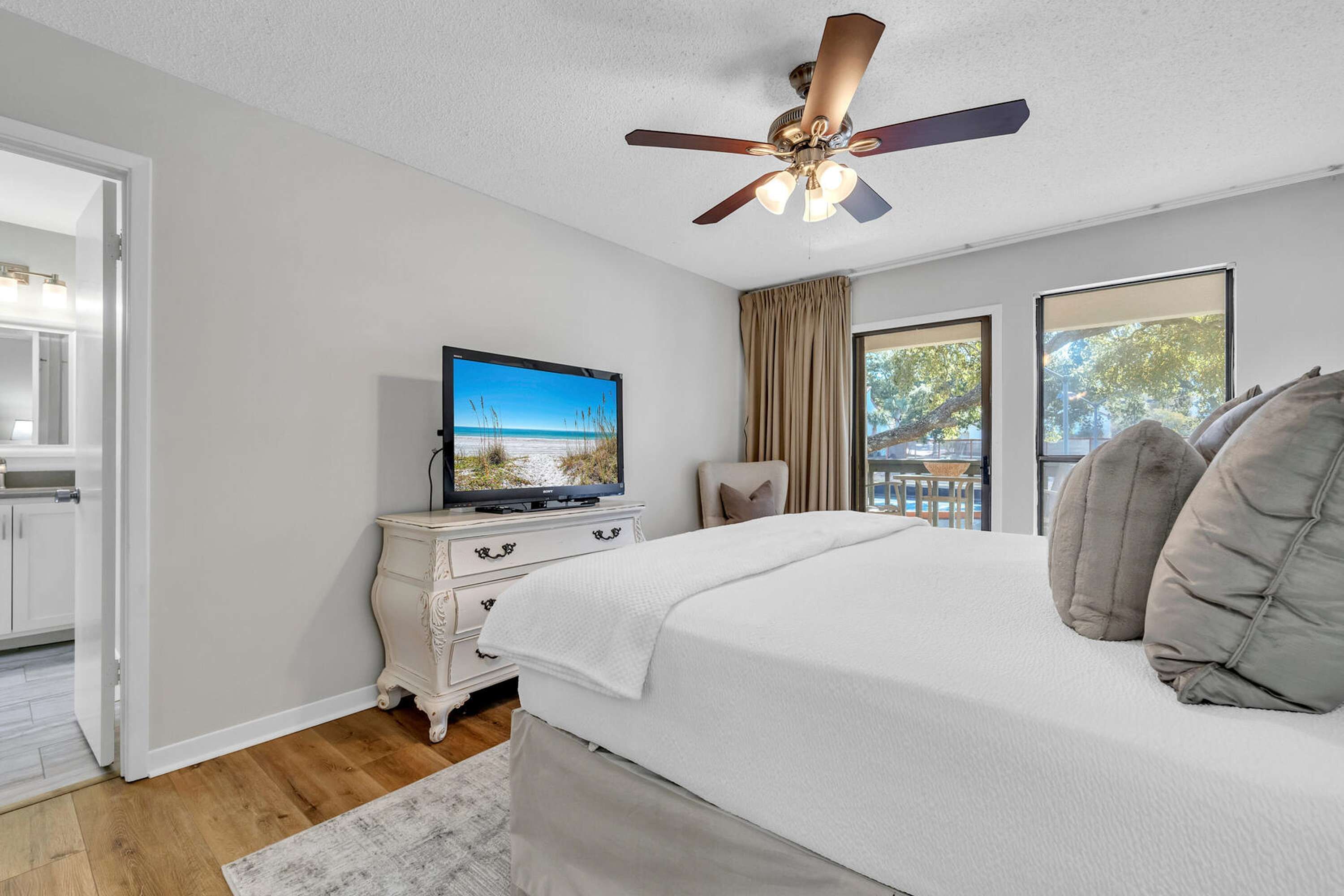 A bedroom with a bed, a ceiling fan, a television, a dresser, and large windows.