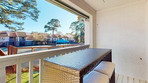 Un balcón con una mesa de mimbre y cojines, con vistas a una zona residencial con casas y un paraguas azul.