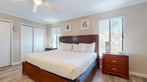 Un dormitorio con cama, ventilador de techo, mesita de noche, cómoda y ventana con persianas.
