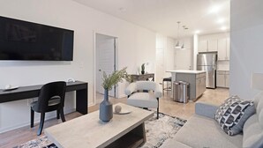 Una sala de estar moderna con un televisor de pantalla plana, un comedor con una silla negra, un sofá gris y una pequeña área de cocina con nevera y armarios.