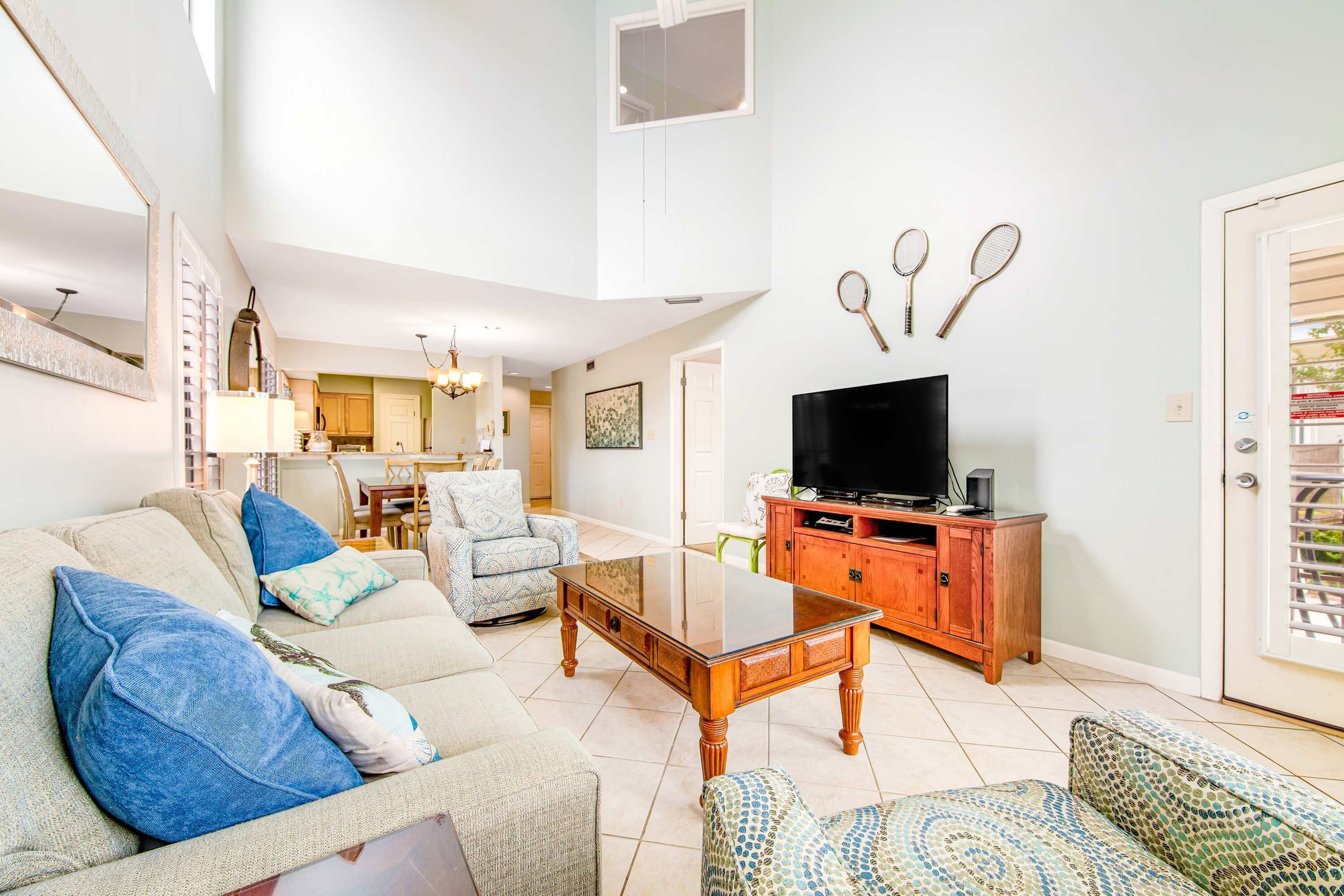 A living room with a sofa, armchair, coffee table, TV stand, and a dining area in the background.