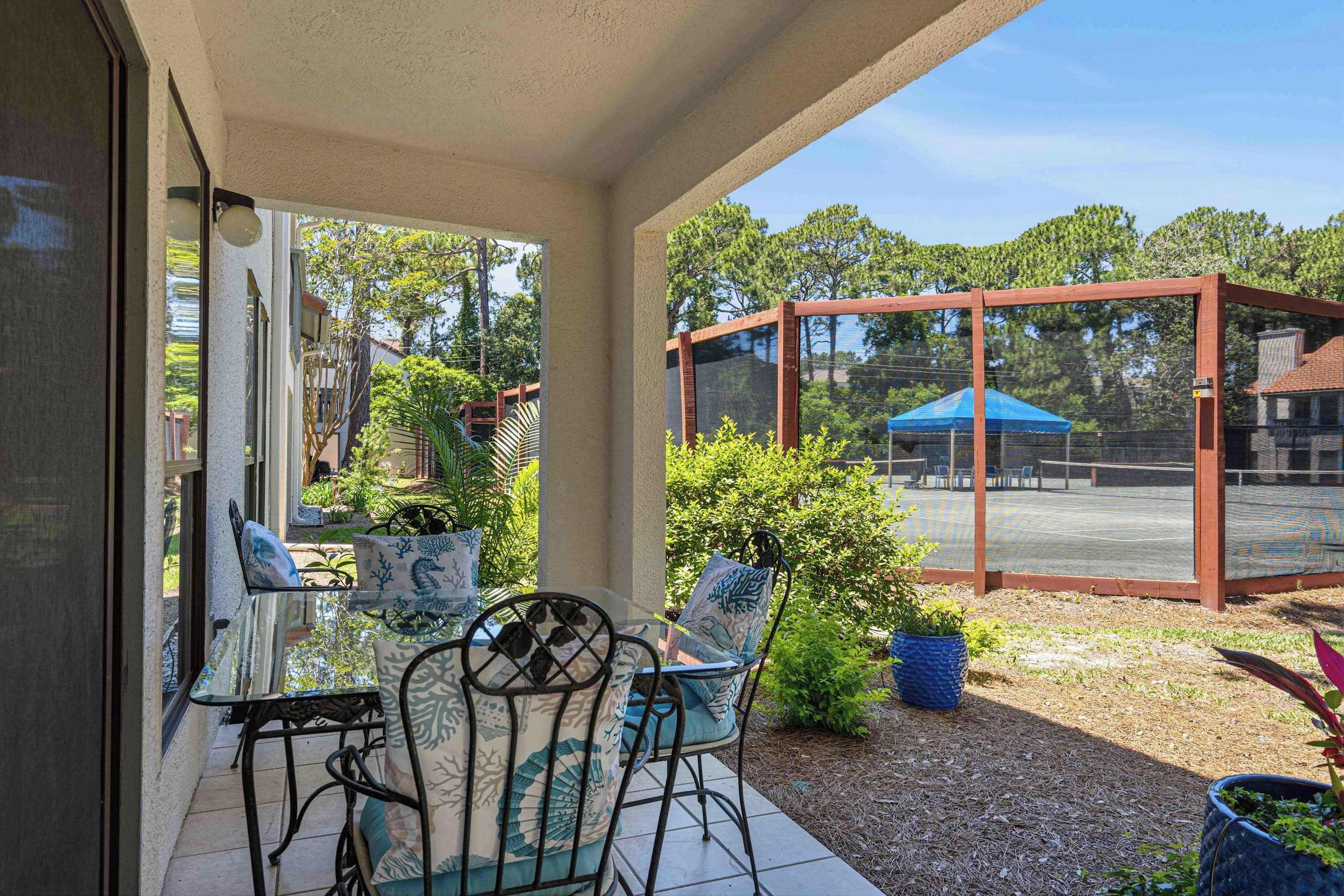 A covered patio area with a glass table and chairs, a wooden pergola, and a view of a tennis court.