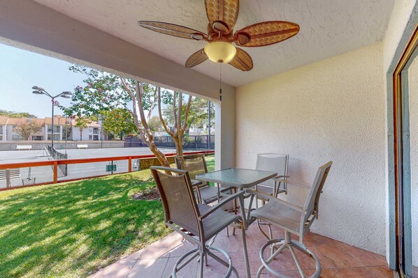 A balcony with a table and chairs, a ceiling fan, and a view of a street and greenery. - Exclusive 2BR tennis condo with beach access (Miramar Beach)