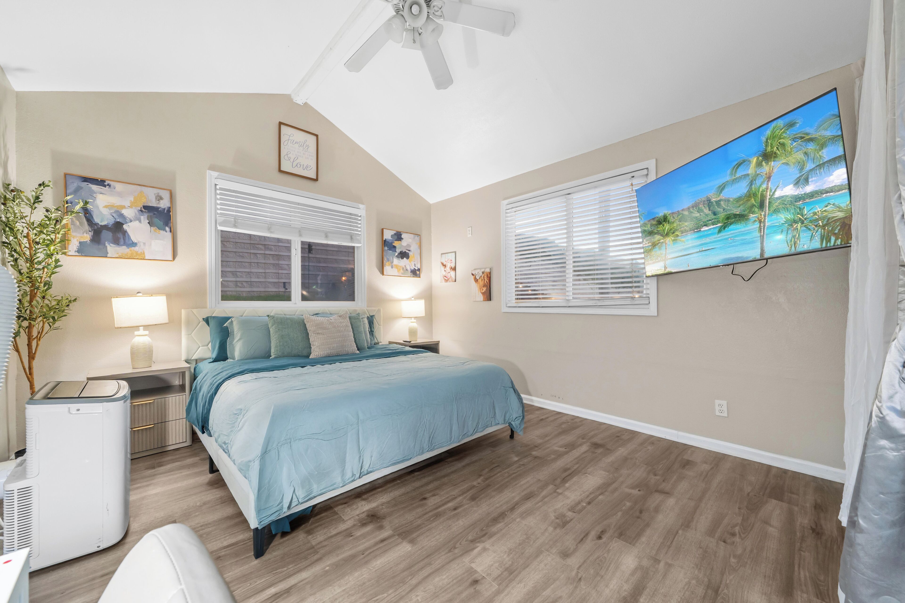 A bedroom with a bed, two bedside tables, a ceiling fan, a large window with blinds, and a wall-mounted TV.