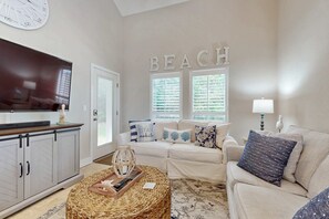 A living room with a sofa, a coffee table, a TV, and a wall with the word 'BEACH' above a window. - Private beach access, tennis retreat, 8 guests (Miramar Beach)