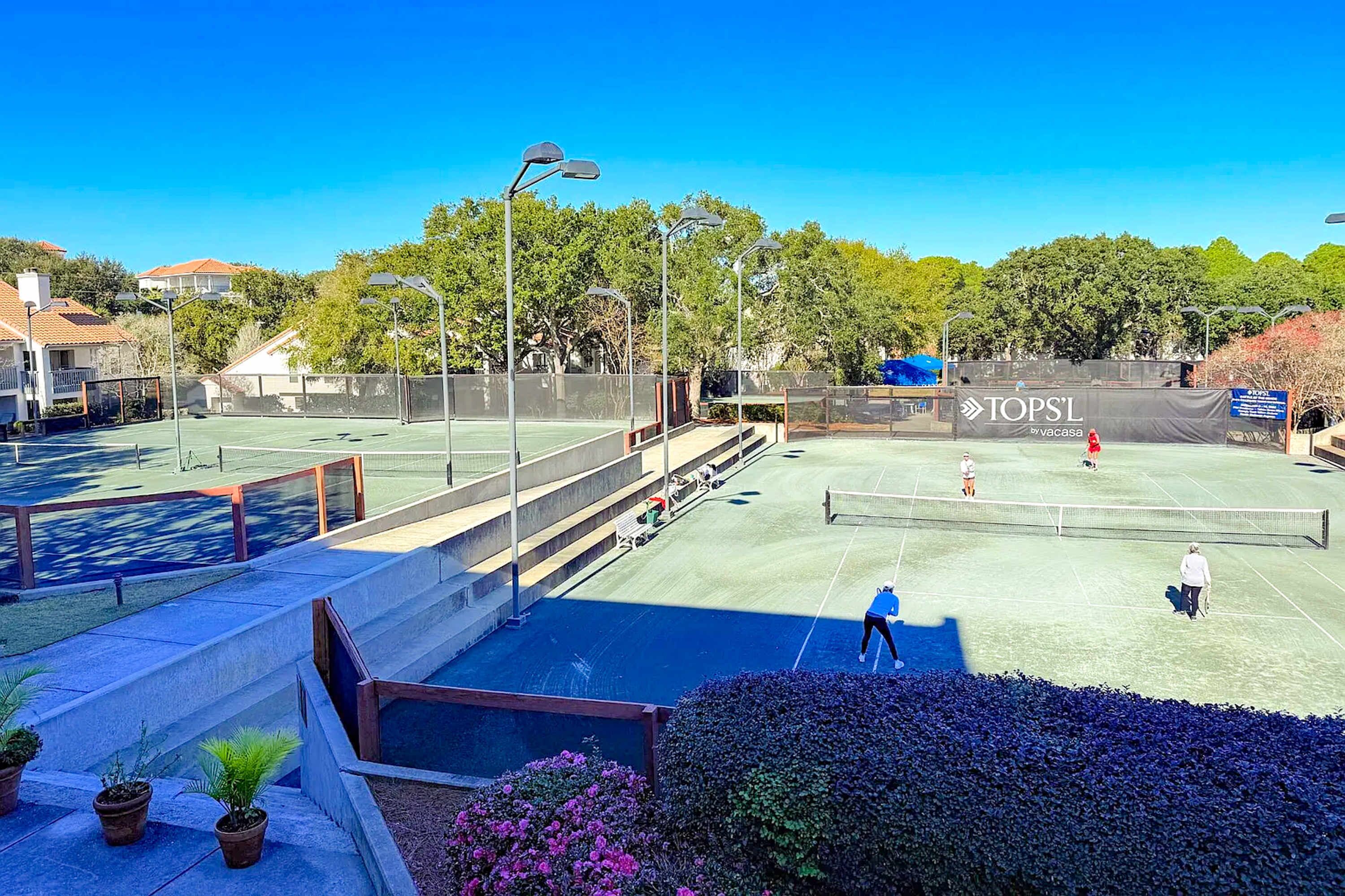 Un court de tennis extérieur, doté d’un filet et entouré d’une clôture, avec une banderole sur laquelle on peut lire le texte « TOPS L ».