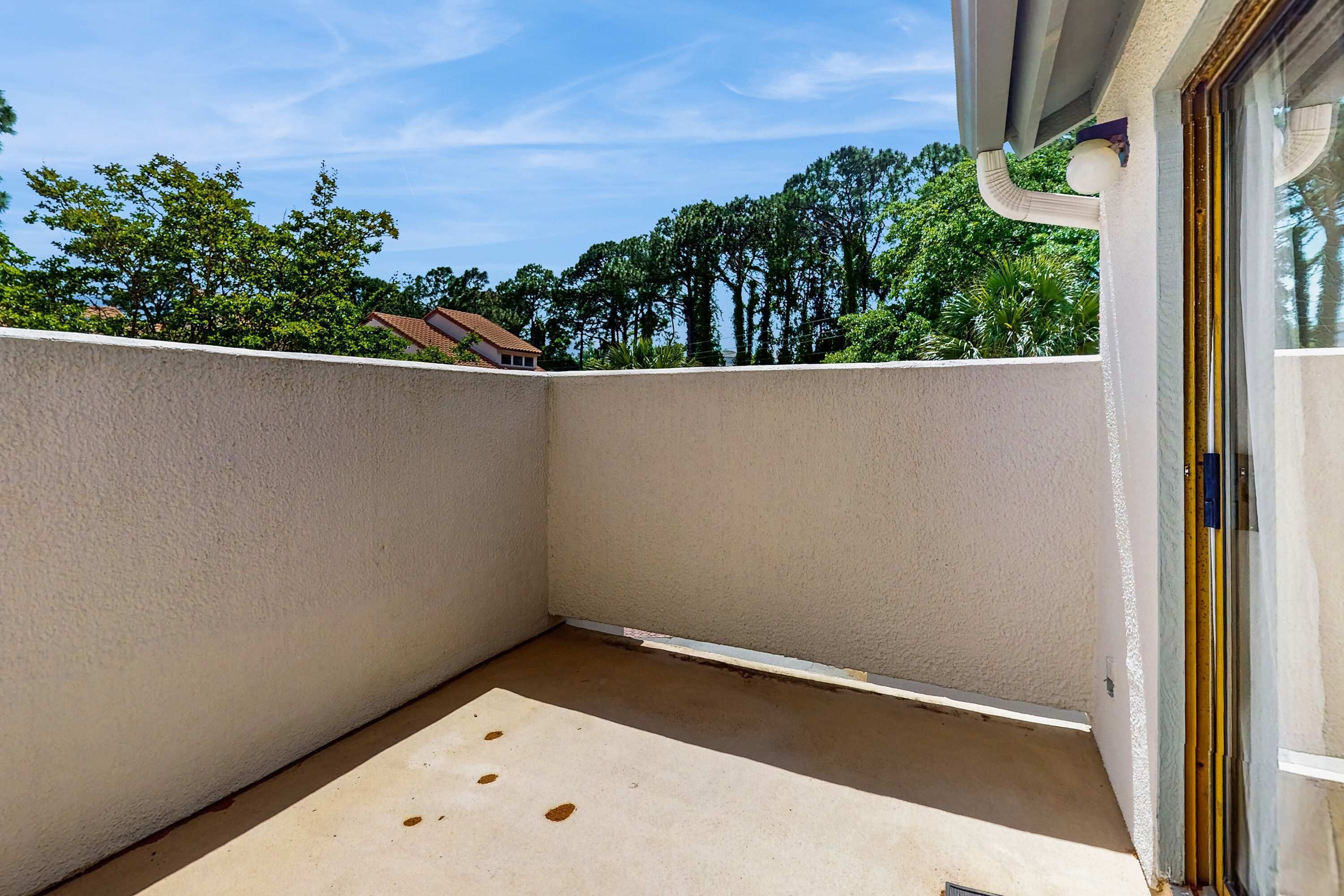 Un balcón con pared blanca, barandilla metálica y una vista despejada de árboles y una casa al fondo.