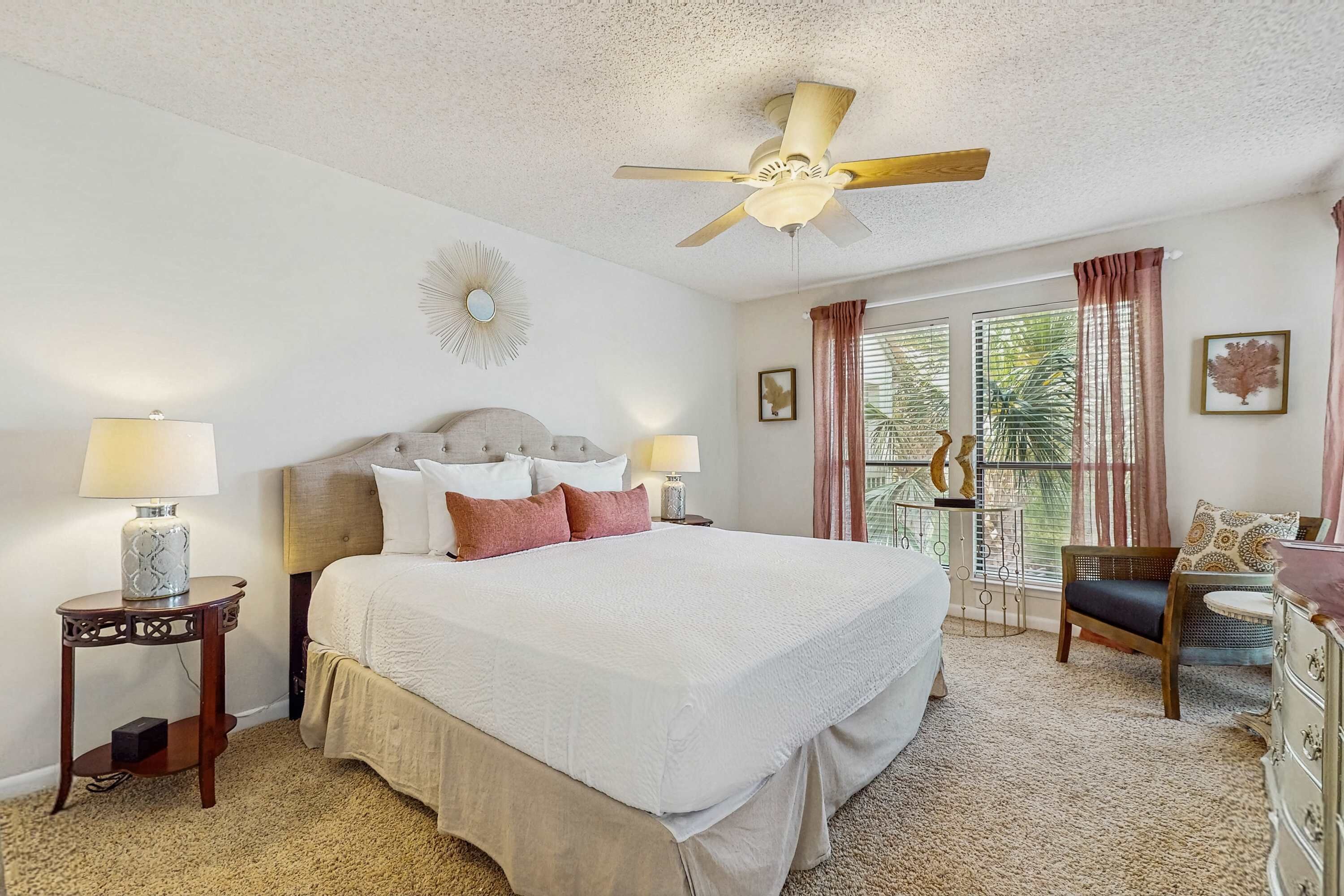 Une chambre d’hôtel avec un grand lit, deux tables de chevet avec des lampes, un ventilateur de plafond, une fenêtre avec des rideaux et une chaise.