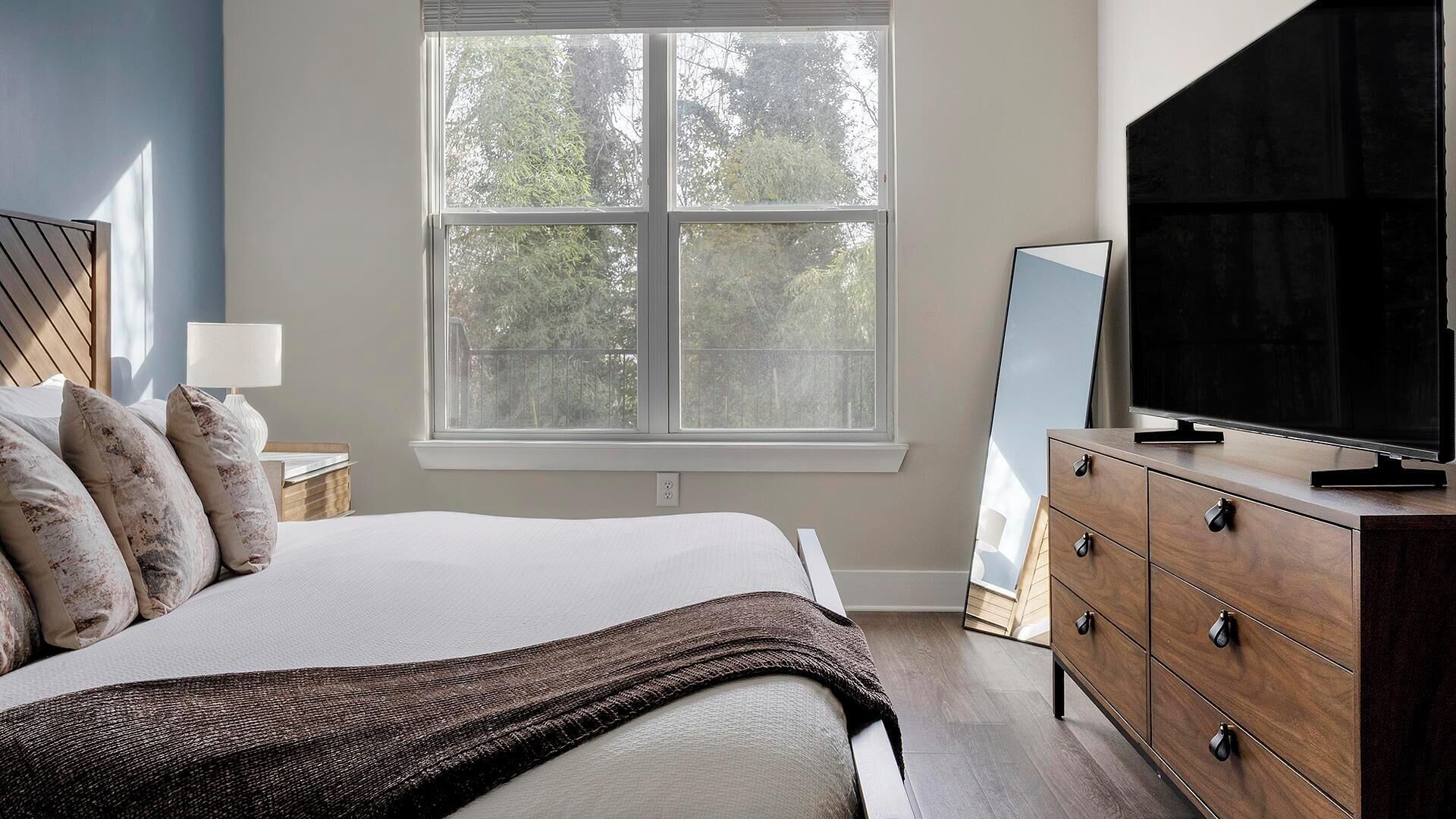 A bedroom with a bed, a dresser, a TV, and a window with a view of trees.