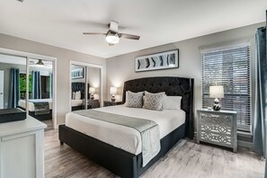 A bedroom with a bed, a ceiling fan, a nightstand, a dresser, a window with blinds, and a door leading to a balcony. - Exclusive 2BR: Beach, Pool & Tennis (Miramar Beach)