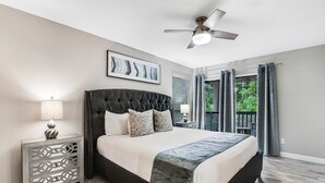 Ein Schlafzimmer mit Bett, Deckenventilator, Nachttisch, Lampe, einem gerahmten Bild und Blick ins Freie.