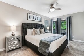 A bedroom with a bed, a ceiling fan, a nightstand, a lamp, a framed picture, and a view of the outdoors. - Exclusive 2BR: Beach, Pool & Tennis (Miramar Beach)