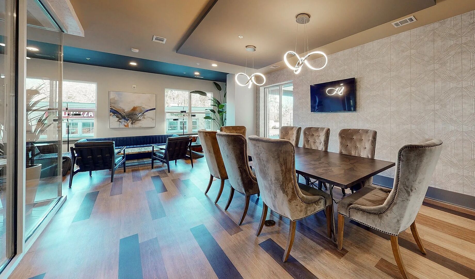 Un espace repas moderne avec une table en bois et des chaises en velours, une grande fenêtre et un téléviseur fixé au mur.