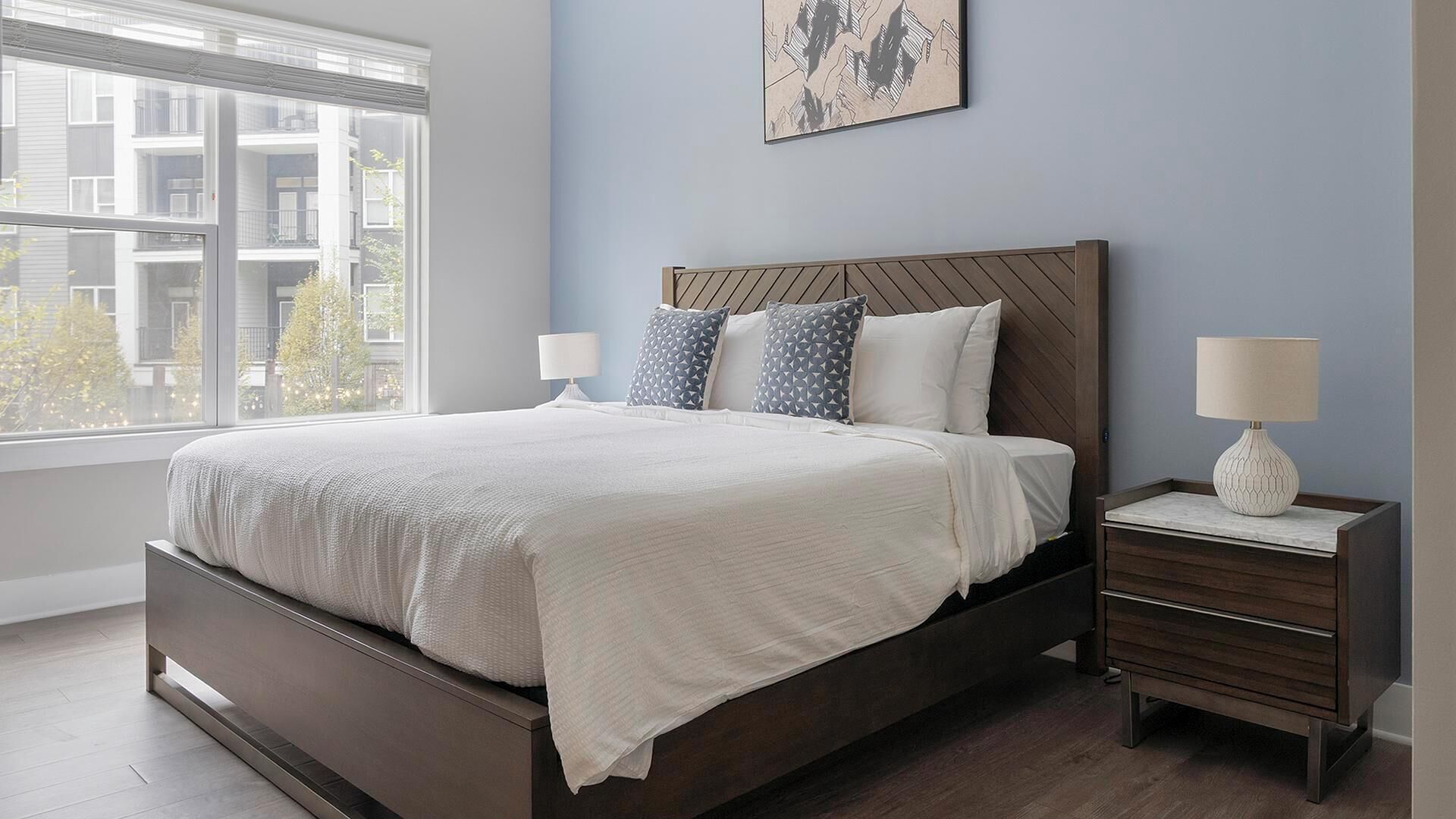 Une chambre moderne avec un grand lit, une table de chevet, une lampe et une fenêtre donnant sur des immeubles.