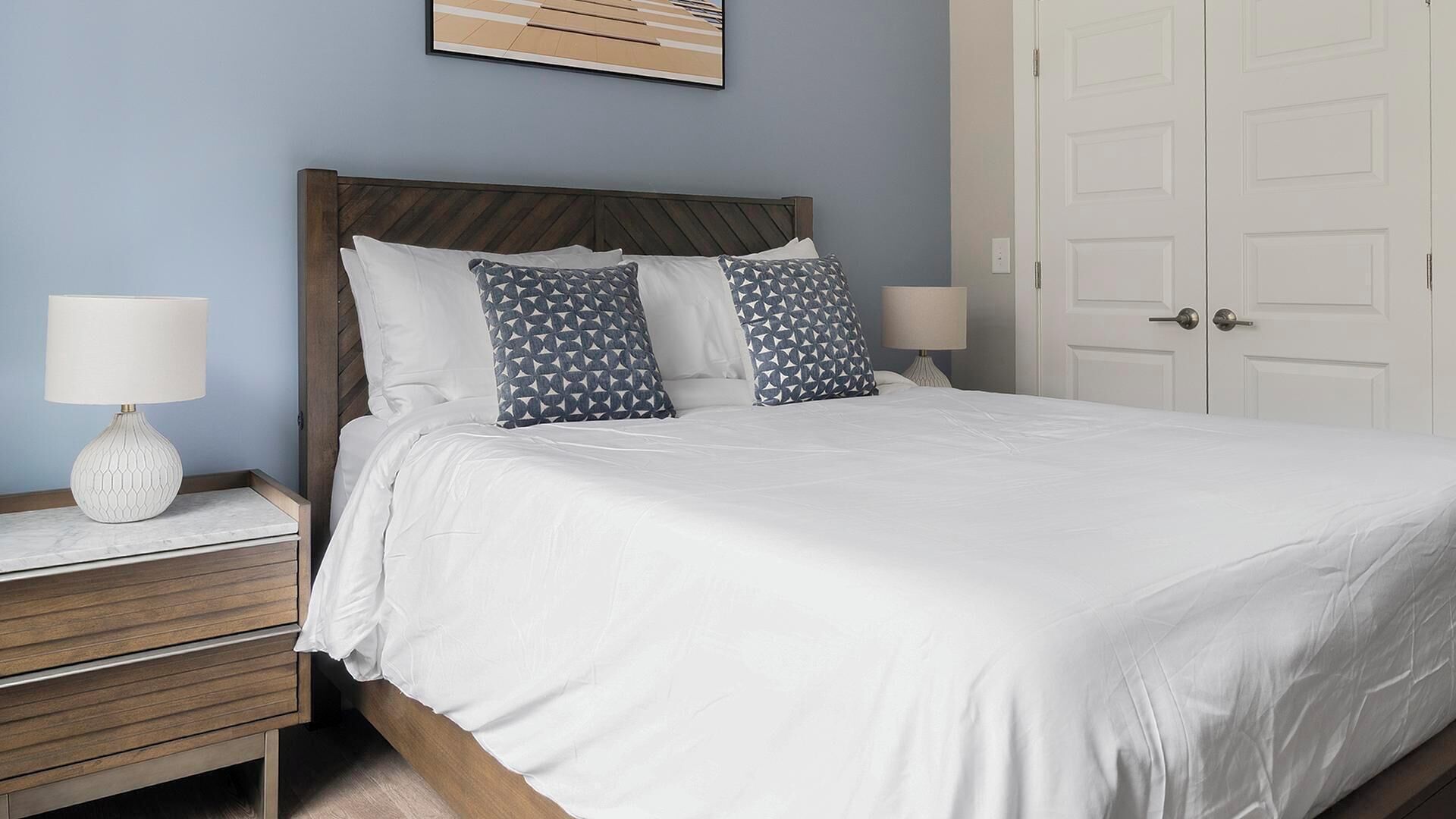 A neatly made bed with white bedding, two patterned pillows, and a wooden headboard. There are two bedside tables with lamps and a framed picture on the wall.
