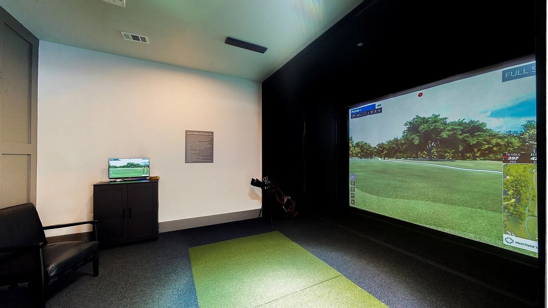 Une pièce dotée d’un grand écran diffusant un match de golf, d’un petit meuble sur lequel est posé un écran, et d’une moquette verte.