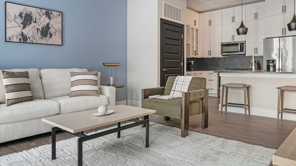 A modern living room with a sofa, armchair, and coffee table, a kitchen with white cabinets and stainless steel appliances, and a blue accent wall. - Landing | Stylish 1BD, Gym, Courtyard (Decatur)