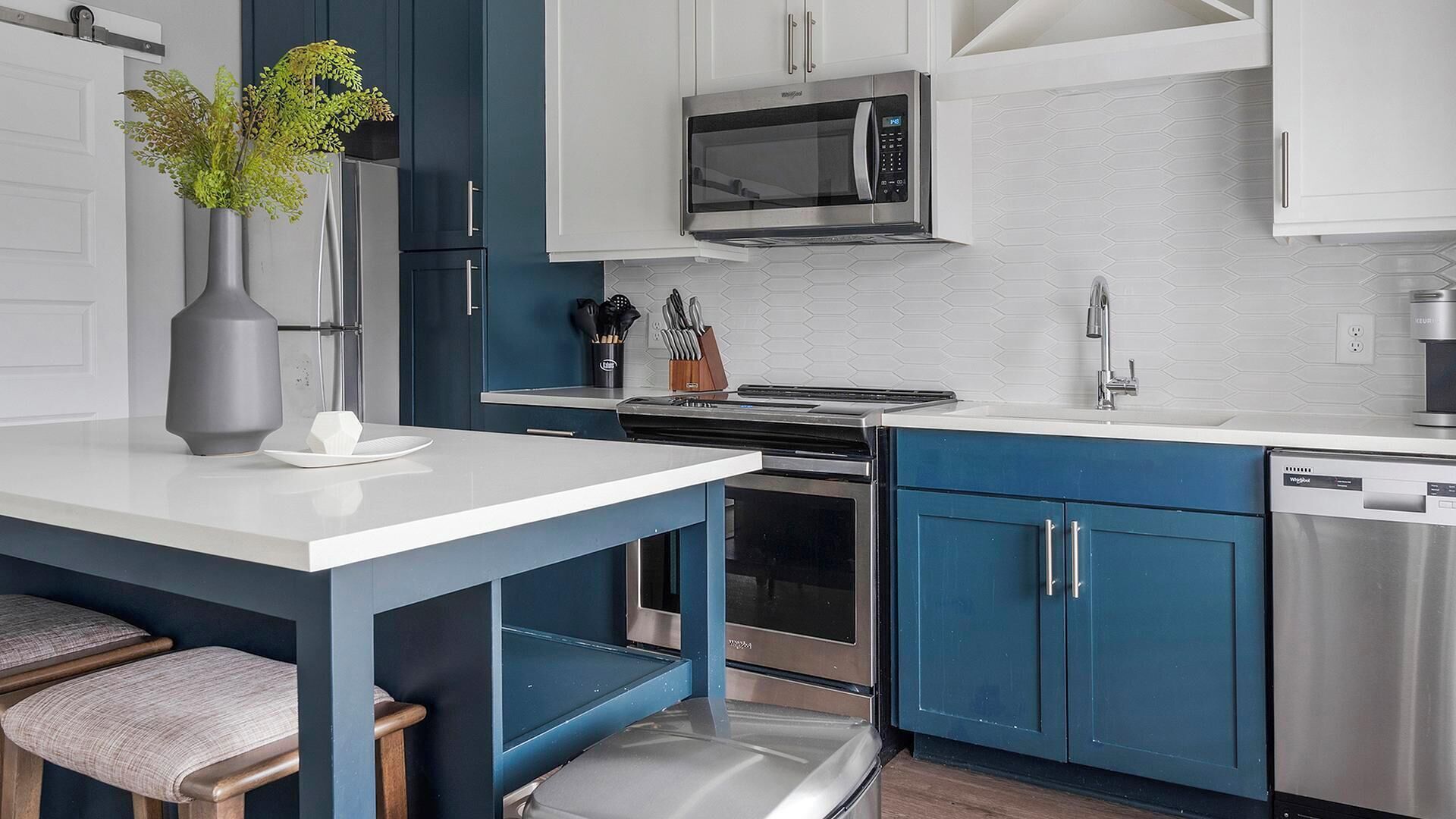 A modern kitchen with a white countertop, blue cabinets, stainless steel appliances, and a vase with green plants.