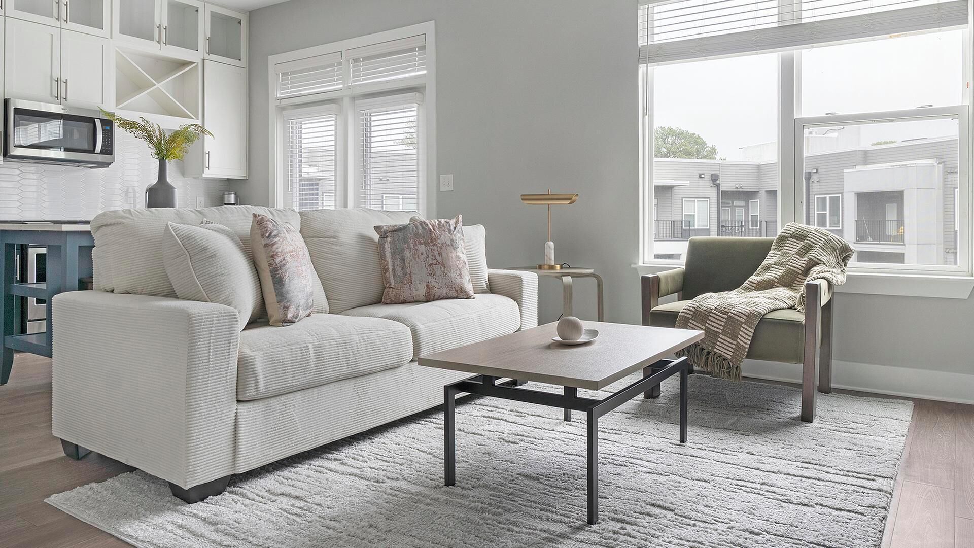 Una sala moderna con un sofá, una mesa de centro y una silla. Hay una zona de cocina con un microondas y armarios.