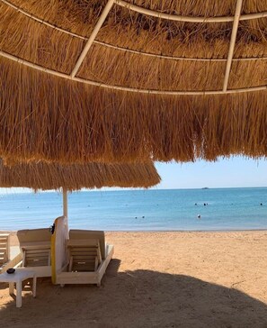 A beach umbrella with a woven design, a lounge chair, and a small table on a sandy beach with clear blue water in the background. -  Luxury Studio, pool, private beach & close to Airport (hurghada)