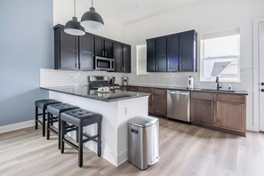 A modern kitchen with dark cabinets, a stainless steel refrigerator, a dishwasher, and a bar area with stools. - Landing | Chic 3BD in Lee's Summit (Lee's Summit)