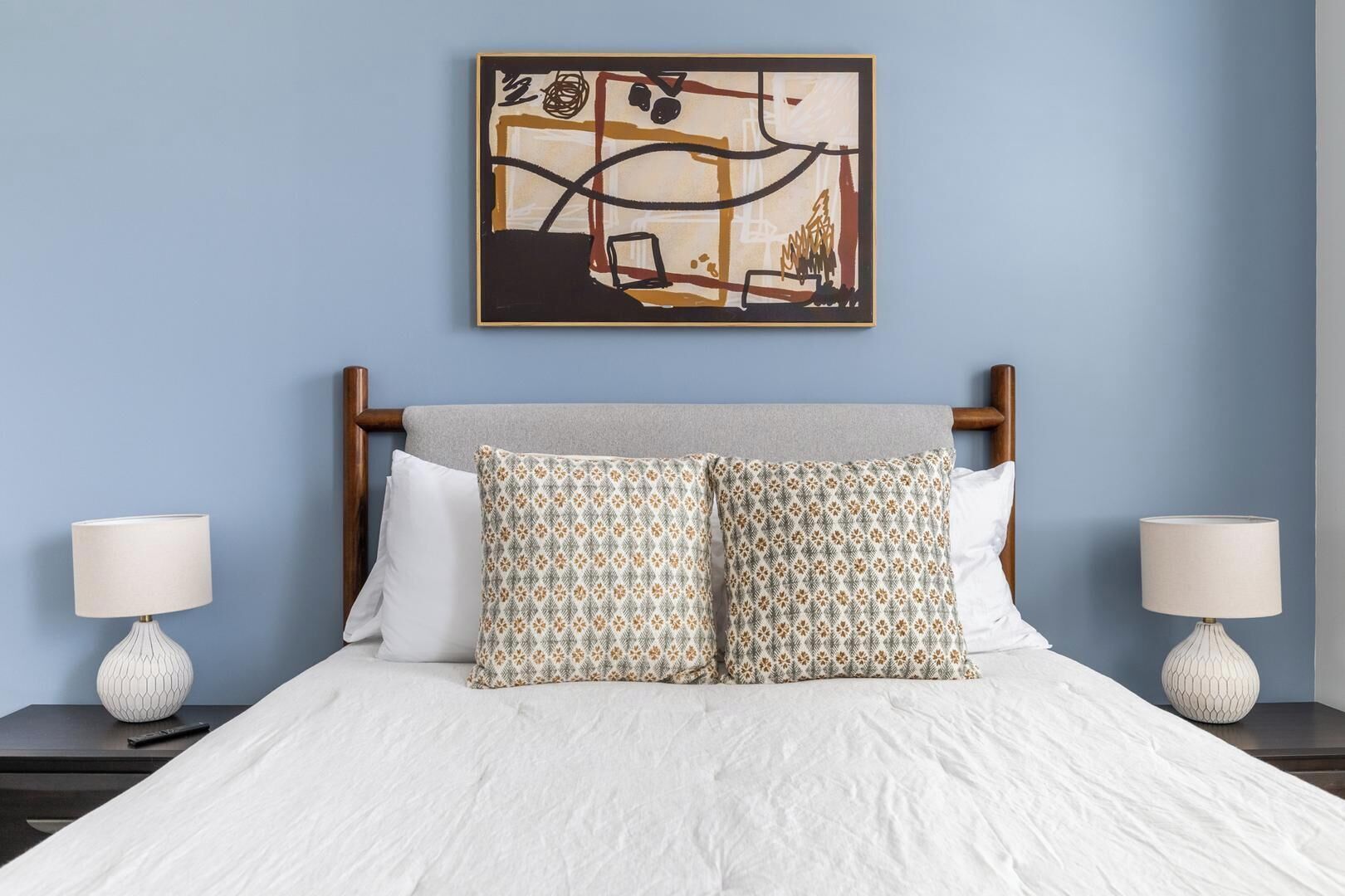 Un lit bien fait, avec des draps blancs, deux coussins à motifs et un tableau abstrait encadré au-dessus.