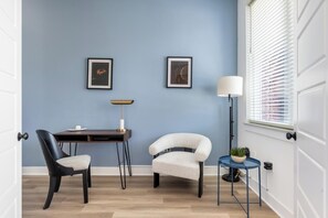 A room with a blue wall, a desk with a chair, a floor lamp, and a window with blinds. - Landing | Stylish 2BD in Lee's Summit (Lee's Summit)