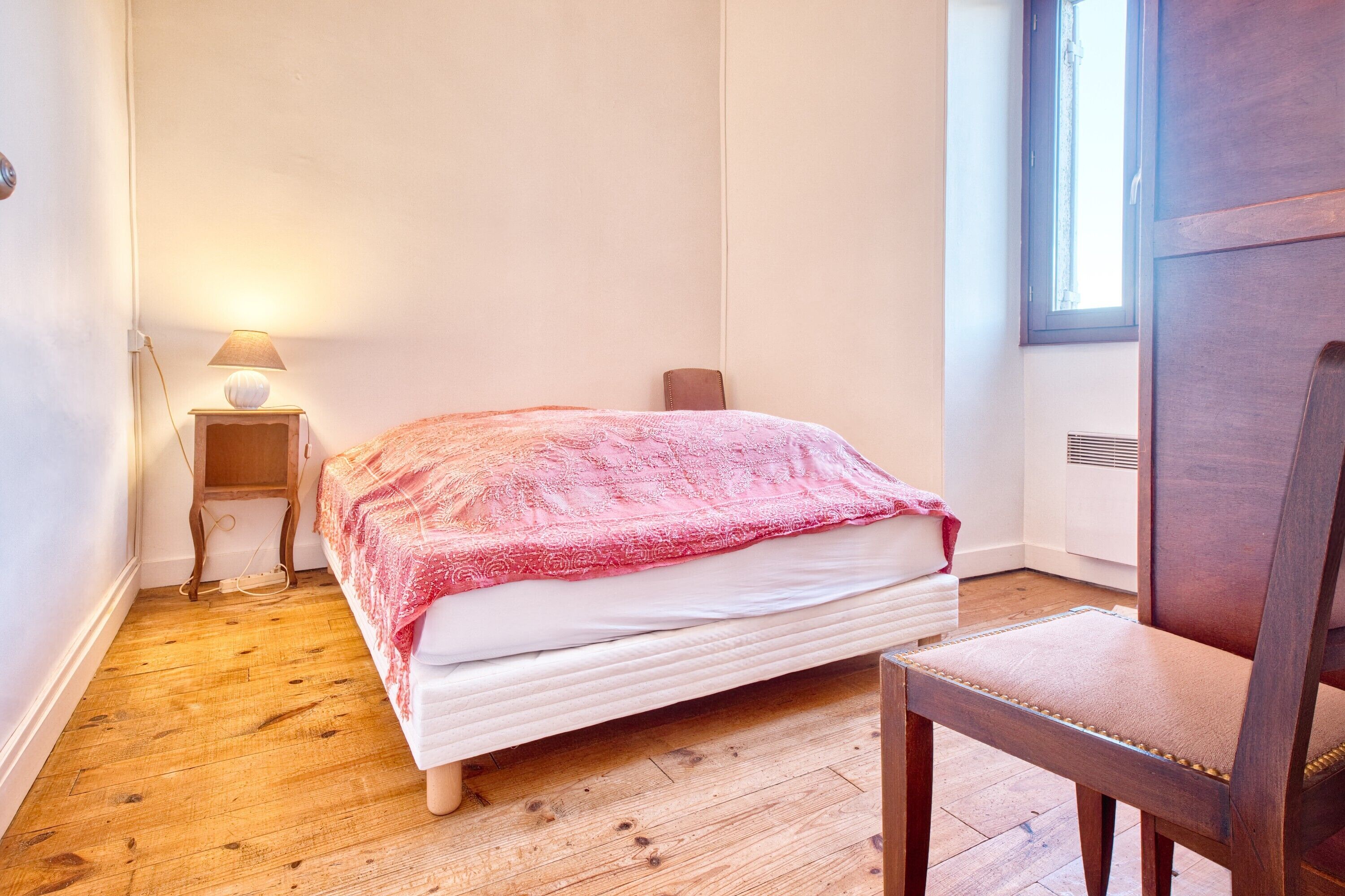 A bedroom with a bed, a chair, a bedside table with a lamp, and a window.