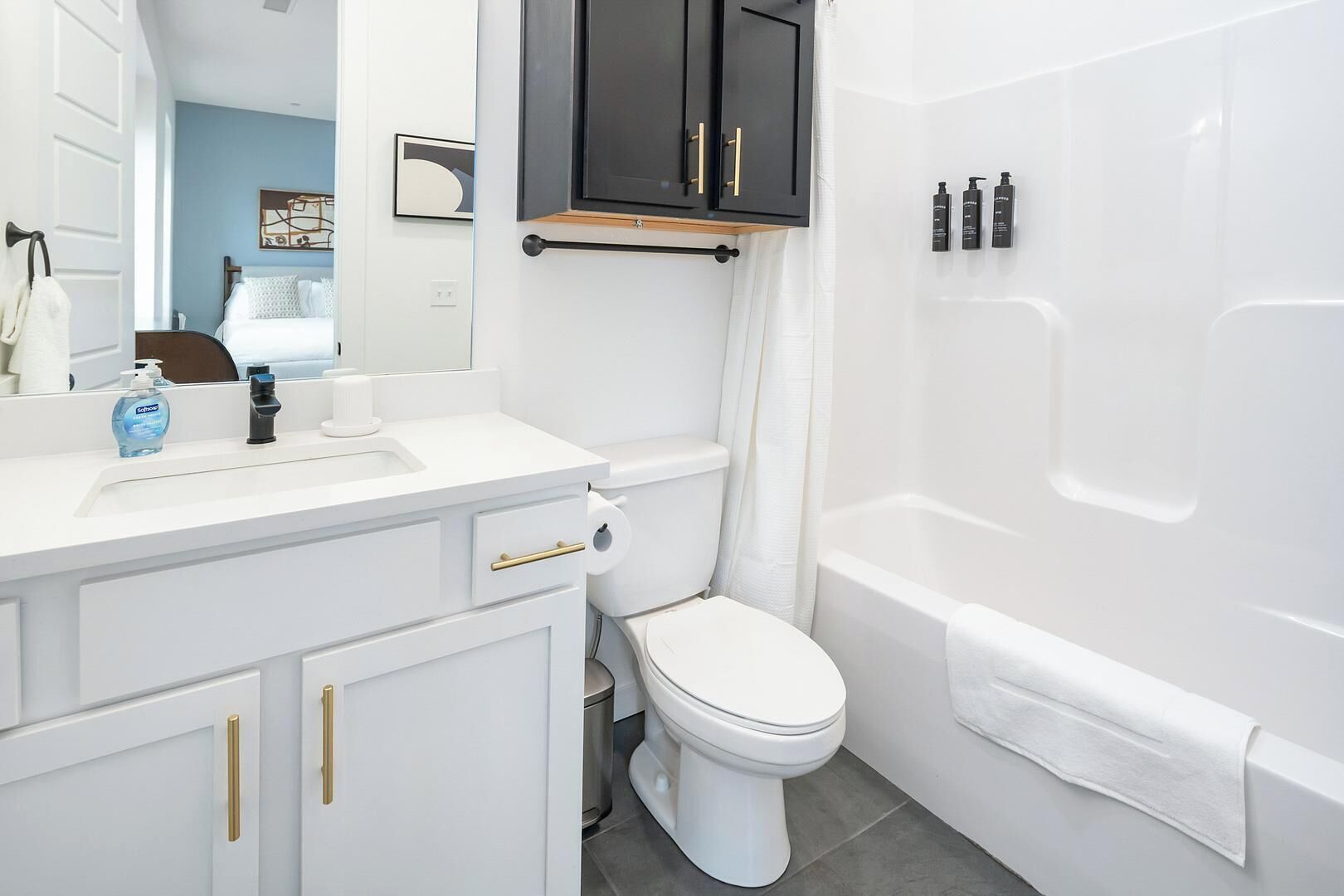 Une salle de bain avec un lavabo, des toilettes et une baignoire, tous de couleur blanche. Un meuble noir est placé au-dessus du lavabo, et une serviette est accrochée au mur.