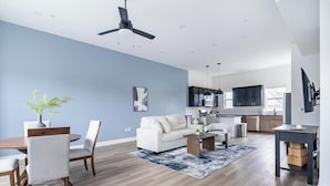Ein modernes Wohnzimmer mit Sofa, Couchtisch und Sesseln, ein Essbereich mit Tisch und Stühlen, eine Küche mit dunklen Schränken und einem Deckenventilator.