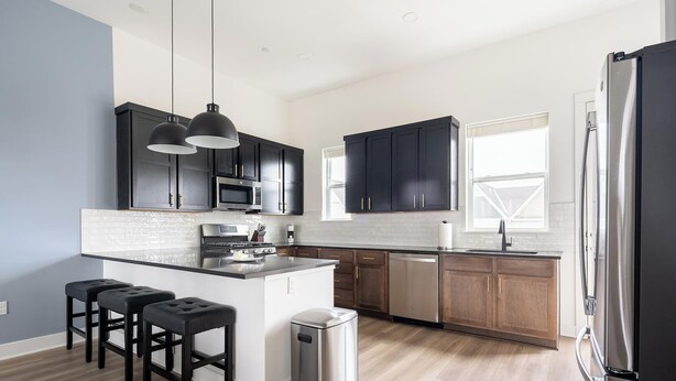 A modern kitchen with dark cabinets, a stainless steel refrigerator, and a central island with bar stools. - Landing | Spectacular 3BD in Lee's Summit (Lee's Summit)