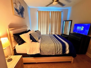 Una habitación de hotel con cama, televisión, ventilador de techo y ventana con cortinas.