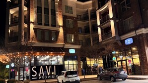 Un immeuble à plusieurs étages, sur lequel on peut voir une grande enseigne « SAVI », indiquant qu’il s’agit d’une boutique de boissons alcoolisées. Des voitures sont garées devant, et l’immeuble possède plusieurs balcons.
