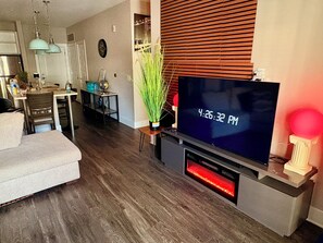 A modern living room with a flat-screen TV, a fireplace, and a dining area in the background.