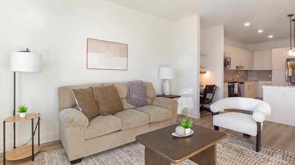 A modern living room with a beige sofa, a coffee table, and a dining area with a chair. - Landing | Spectacular 1BD, Gym, Clubhouse (Longmont)