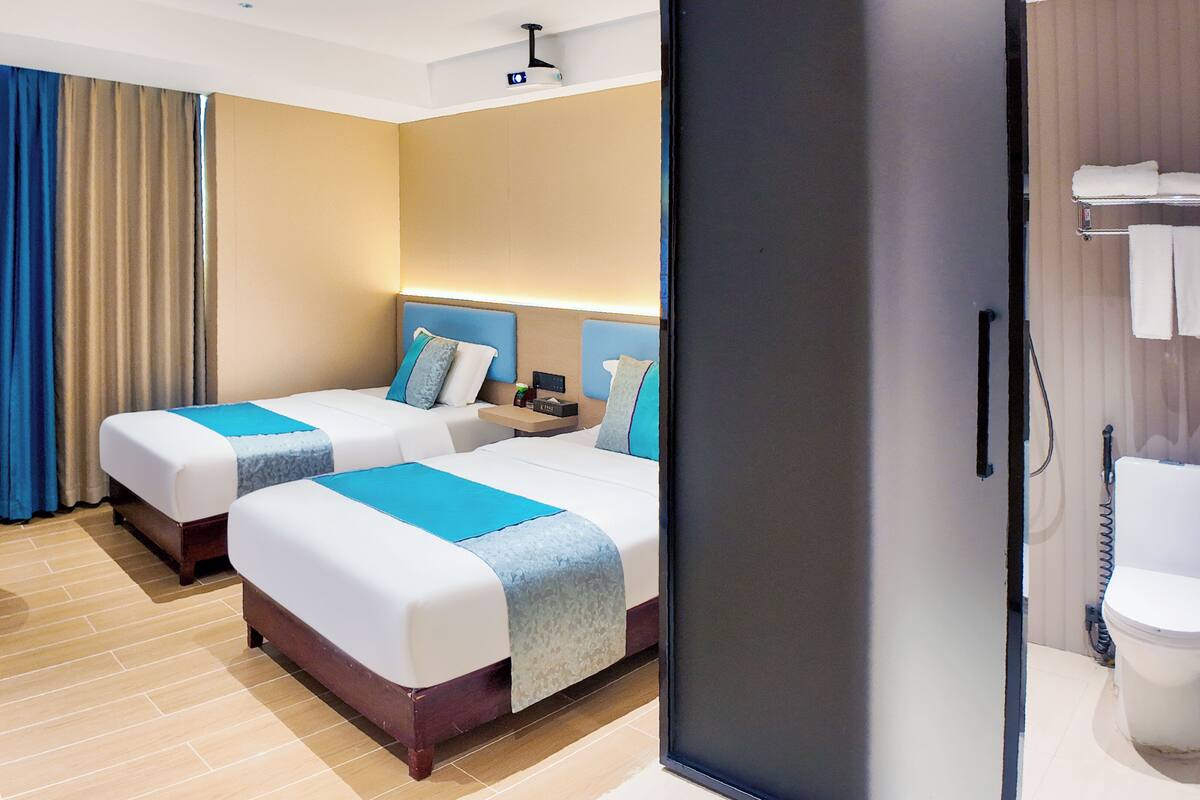 A hotel room with two beds, a bedside table, a lamp, a bathroom with a toilet and shower, and a curtain.