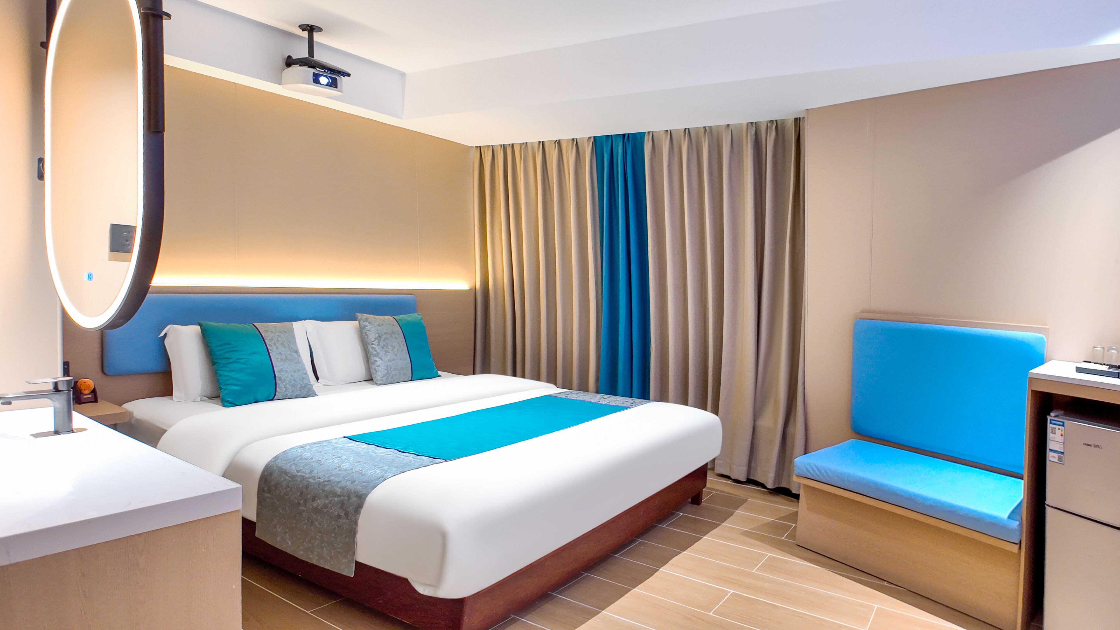Ein Hotelzimmer mit einem großen Bett, einem Nachttisch, einem Spiegel, einem blauen, gepolsterten Sessel und einem kleinen Schreibtisch mit Lampe.