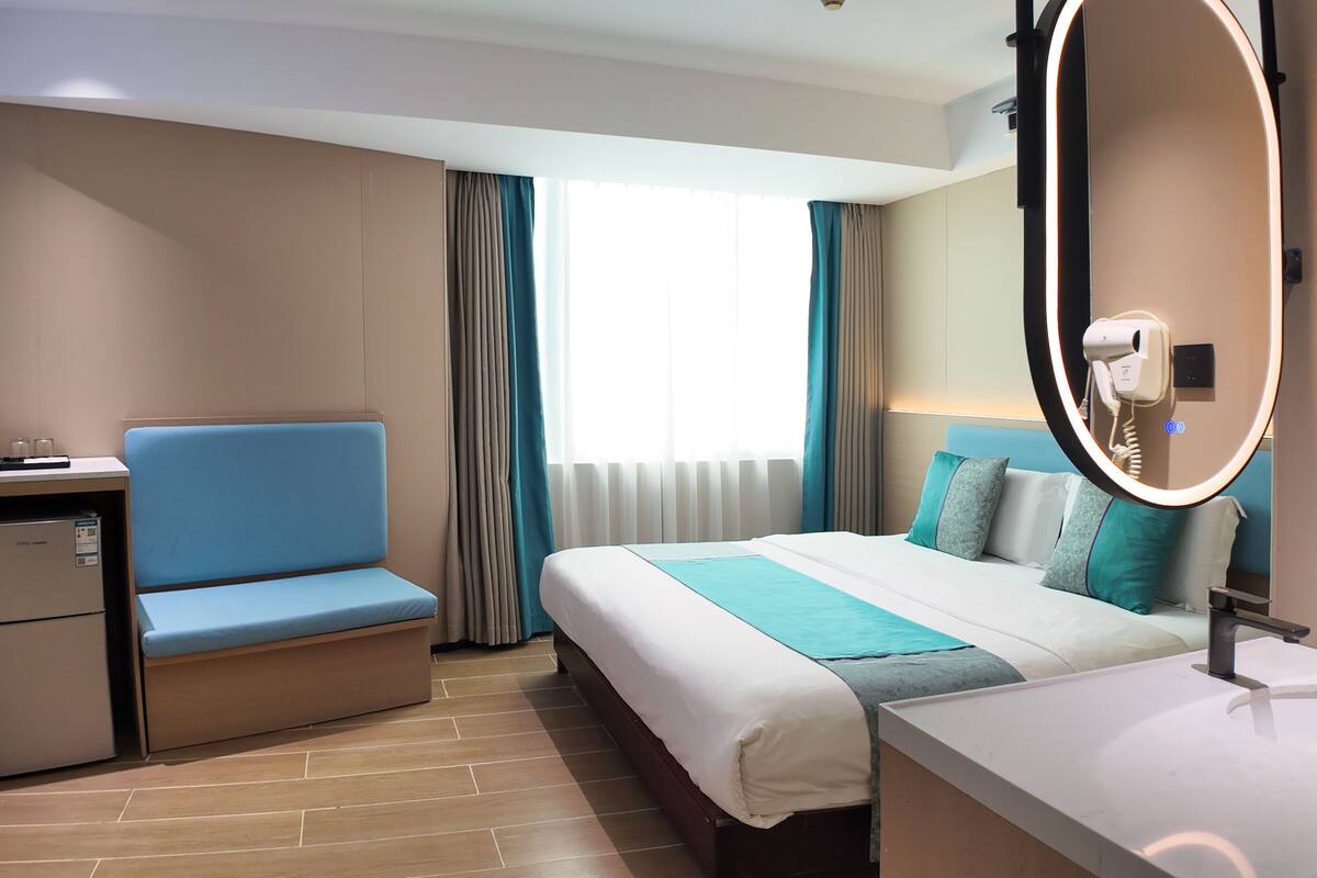 A hotel room with a bed, a desk with a chair, a small refrigerator, a mirror, and a sink.