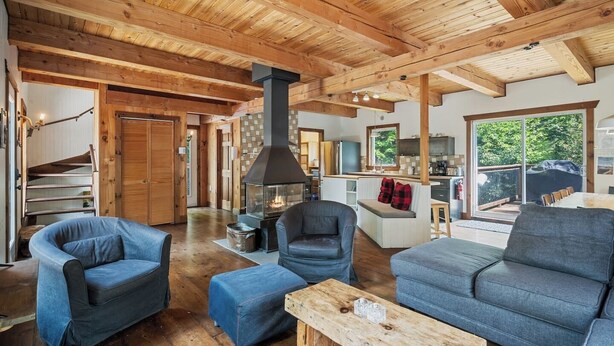 A cozy cabin interior with a central wood-burning stove, a dining area, and a large window overlooking a deck.