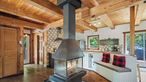 A rustic cabin interior with a central wood-burning stove, a kitchen area, and a seating area with a view of the outdoors.
